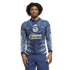 A man models a blue long-sleeved Club América soccer jersey with ornate light blue patterns and "Caliente" logo, wearing blue jeans against a black background.