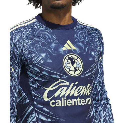 Close-up of a navy blue Club América soccer jersey with a light blue eagle pattern, yellow Adidas logo, and "Caliente" sponsor logo.