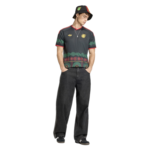 A man in a black Adidas Jamaica jersey with colorful patterns, matching bucket hat, and wide-leg dark jeans stands against a black background.