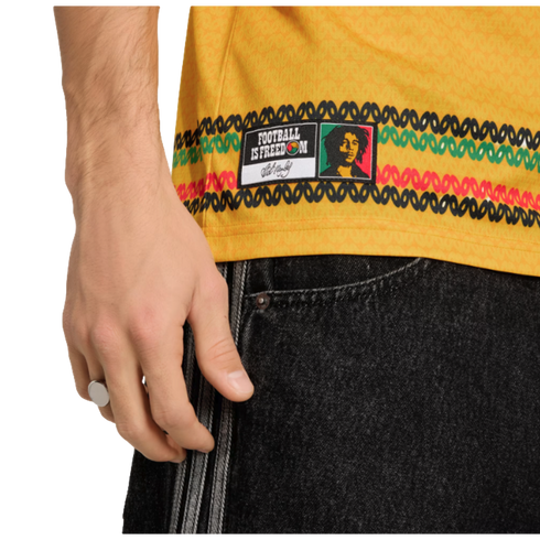 Close-up of a yellow jersey with a Bob Marley "Football is Freedom" patch and knit-patterned stripes, worn with black striped denim. A silver ring is visible on the person's hand.