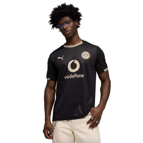 A man with glasses and curly hair wears a black and gold Borussia Dortmund soccer jersey with a Vodafone logo, posing against a black background.