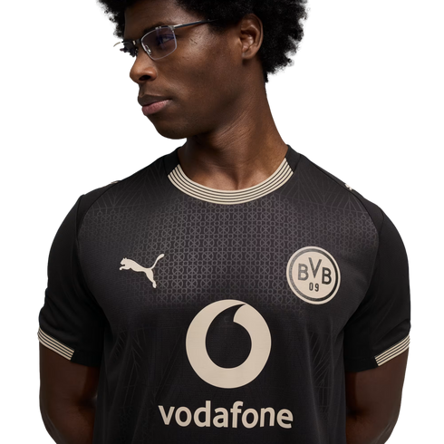 A man wearing glasses and a black Puma football jersey featuring the Borussia Dortmund (BVB) crest and a Vodafone sponsor logo, set against a black background.