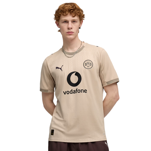 A man with curly red hair wearing a beige Borussia Dortmund soccer jersey with a Vodafone logo, posing against a black background.