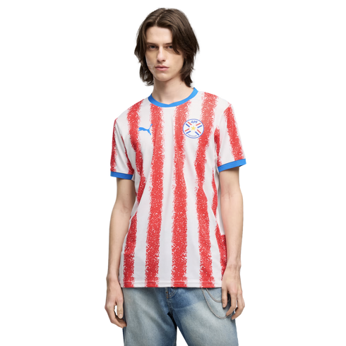 A man wearing a white and red vertically striped Paraguay national team soccer jersey and blue jeans, standing against a black background.