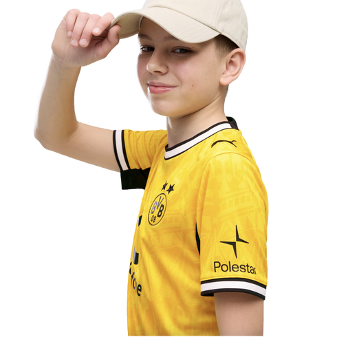 A smiling boy in a yellow Borussia Dortmund soccer jersey and beige baseball cap, touching the brim of his hat against a black background.