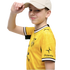 A smiling boy in a yellow Borussia Dortmund soccer jersey and beige baseball cap, touching the brim of his hat against a black background.