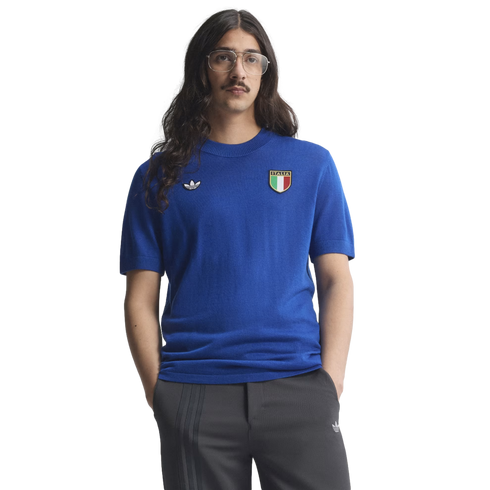 A man with long dark hair, glasses, and a mustache wearing a blue Adidas Italia knit shirt and grey trousers against a white background.