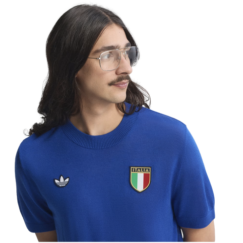 A man with long dark hair and a mustache wears a blue knit Adidas sweater with an Italia crest, looking to the side.