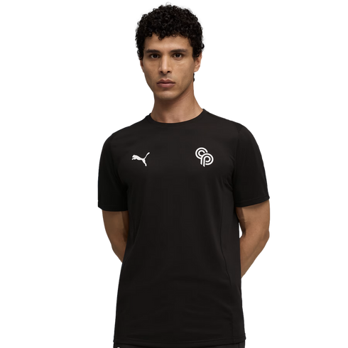 Man with curly dark hair in a black Puma tee with white logos on the chest, posing with hands behind his back against a plain black background.