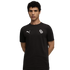 Man with curly dark hair in a black Puma tee with white logos on the chest, posing with hands behind his back against a plain black background.