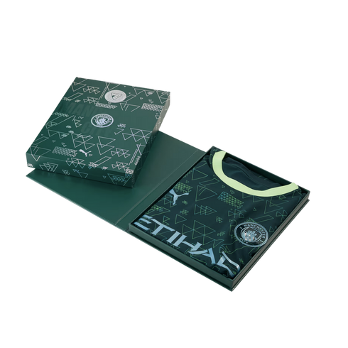 A dark green Manchester City football jersey with a geometric pattern and lime green collar, presented in its matching collector's box against a black background.