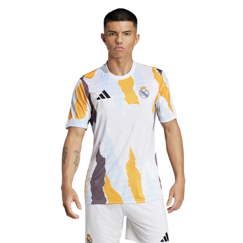A model wearing a Real Madrid pre-match jersey and white shorts against a black background. The white shirt features a bold, jagged abstract pattern in yellow, grey, and light blue, with the black Adidas logo and Real Madrid club crest on the chest.