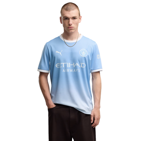A man wearing a light blue Manchester City soccer jersey and dark pants, posing against a plain white background.