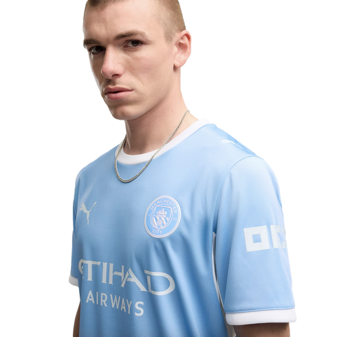 A young man with a short buzz cut wearing a light blue Manchester City soccer jersey and a silver chain poses against a white background.