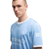 A young man with a short buzz cut wearing a light blue Manchester City soccer jersey and a silver chain poses against a white background.