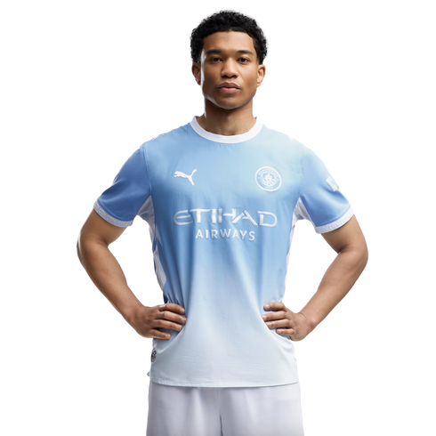 A young man wearing a light blue Manchester City soccer kit poses with his hands on his hips against a white background.