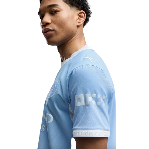 Side profile of a young man with curly hair wearing a light blue Manchester City football jersey against a white background.