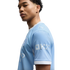 Side profile of a young man with curly hair wearing a light blue Manchester City football jersey against a white background.