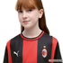 A young girl with long red hair and freckles wears a red and black striped AC Milan football jersey.