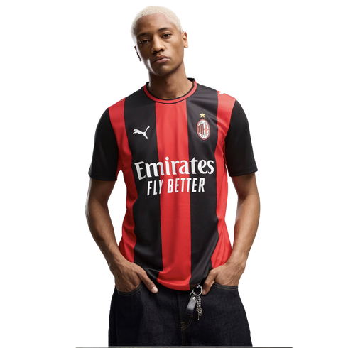 A man with short blonde hair wears a red and black AC Milan jersey and dark jeans, posing with hands in his pockets against a white background.