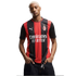 A man with short blonde hair wears a red and black AC Milan jersey and dark jeans, posing with hands in his pockets against a white background.