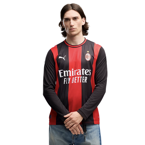 A man with mid-length dark hair wearing an AC Milan long-sleeved soccer jersey and blue jeans, standing against a plain white background.