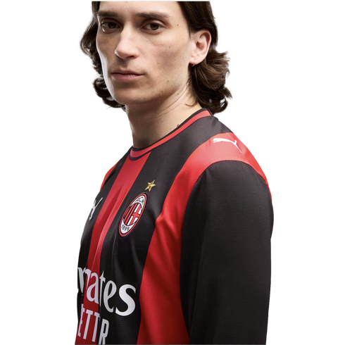 A man with long dark hair wearing a red and black striped AC Milan jersey against a white background.