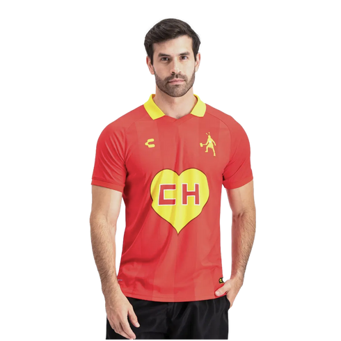 A man with a beard wearing a red jersey with a yellow collar and a large yellow heart centered with red "CH" letters. He stands against a white background with one hand in his pocket.